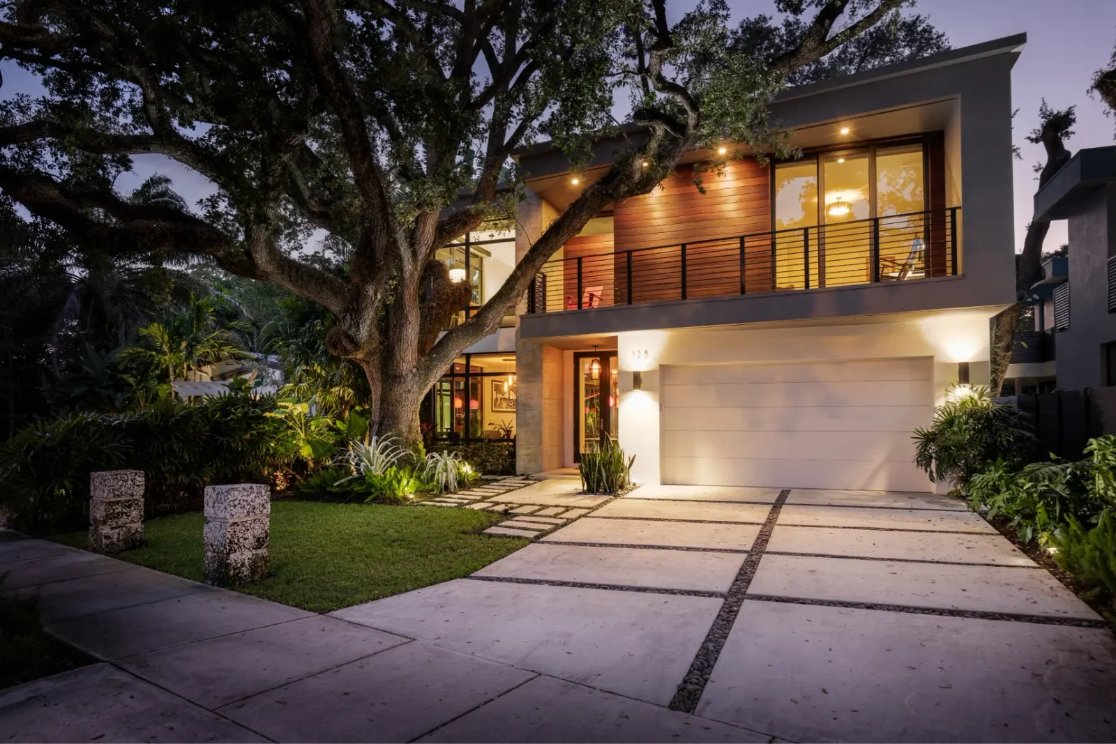 Contemporary two-story home with large windows and lush landscaping, illuminated at dusk.